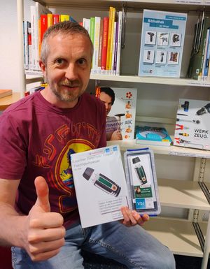 Bibliotheksmitarbeiter Marko Schönwelski hält den Feuchtigkeitsmesser in der Verleihbox vor dem Regal mit den Medien zum Heimwerken in der Hand und macht die Daumen-hoch-Geste
