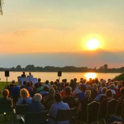 Bild von der Lesung 2019; Zuhörer sitzen mit dem Gesicht zur Bühne und den Autoren darauf am Inselseestrand, dahinter spiegelt sich die untergehende Sonne im See