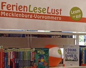 Banner mit dem Schriftzug FerienLeseLust Mecklenburg-Vorpommern und dem runden grünen Logo mit dem Schriftzug "Lesen tut gut", darunter eine Reihe ausleihbare Bücher für den Ferienleseclub und ein aufgeblasener Wasserball mit dem gleichen Logo