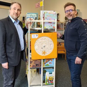 Tilmann Wesolowski und Steffen Büchner präsentieren das neue Wandspiel im Kinderraum, das an der Seite eines Buchregals angebracht ist.