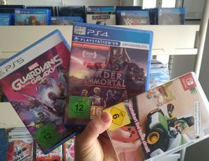 Drei Hüllen von Konsolenspielen werden vor dem Spiele-Regal der Bibliothek hochgehalten