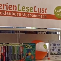 Banner mit dem Schriftzug FerienLeseLust Mecklenburg-Vorpommern und dem runden grünen Logo mit dem Schriftzug "Lesen tut gut", darunter eine Reihe ausleihbare Bücher für den Ferienleseclub und ein aufgeblasener Wasserball mit dem gleichen Logo