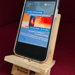 Ein Smartphone steht auf einem der Handyhalter, die man mit Quizrallye gewinnen kann, und auf dem Display ist das Plakat zur Veranstaltung zu sehen