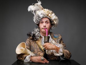 Philipp Weber mit Barocker Perrücke und Wams, im Arm ein Megaphon und an einen rosa Strohhalm aus einem Kupferbecher trinkend; (c) S. Büttner