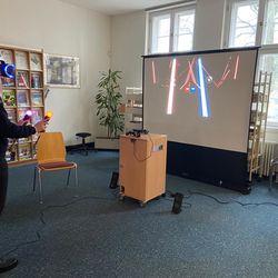 Person mit aufgesetzter VR-Brille und Move-Controllern in der Hand vor der VR-Station der Bibliothek und der aufgebauten Leinwand, auf der die Zuschauer das Bild aus der Brille übertragen bekommen