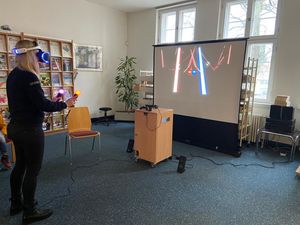 Person mit aufgesetzter VR-Brille und Move-Controllern in der Hand vor der VR-Station der Bibliothek und der aufgebauten Leinwand, auf der die Zuschauer das Bild aus der Brille übertragen bekommen