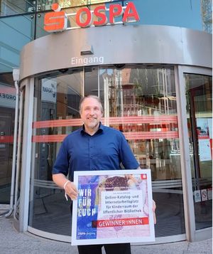 Bibliotheksverantwortlicher Tilmann Wesolowski mit der Förderurkunde in der Hand vor dem Eingang der OSPA-Zentrale in Rostock